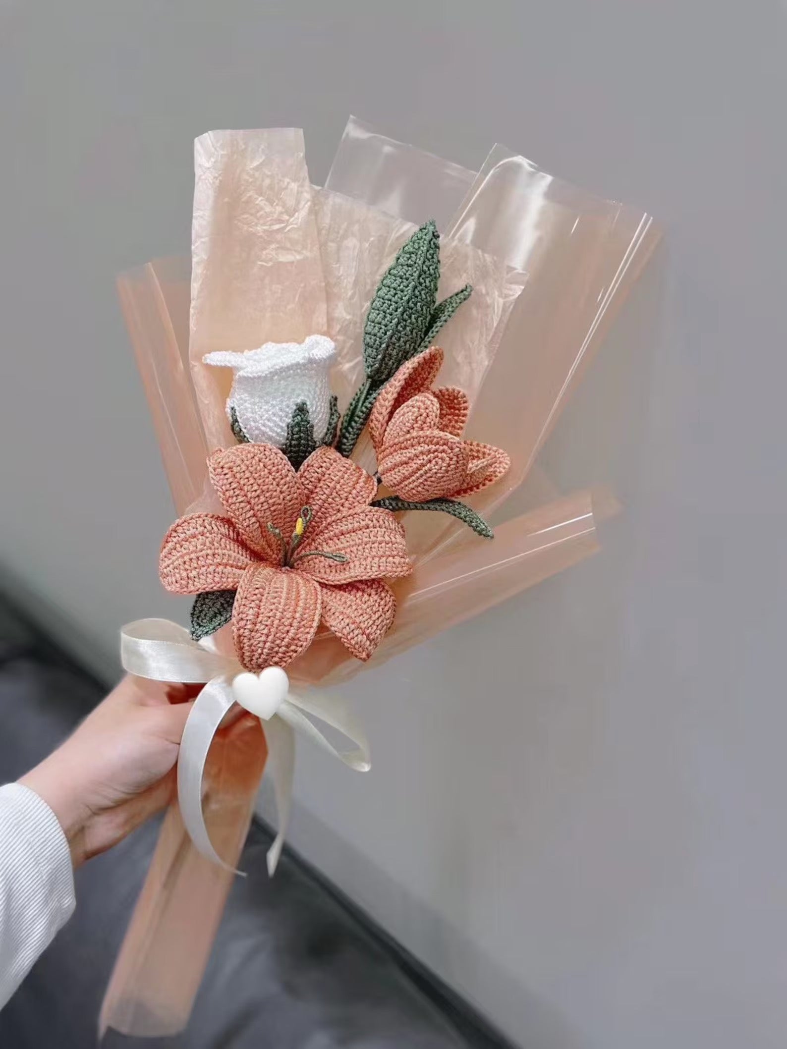 A crochet lily bouquet with roses, tulips, and green leaves in shades of white and orange, tied with ribbons.